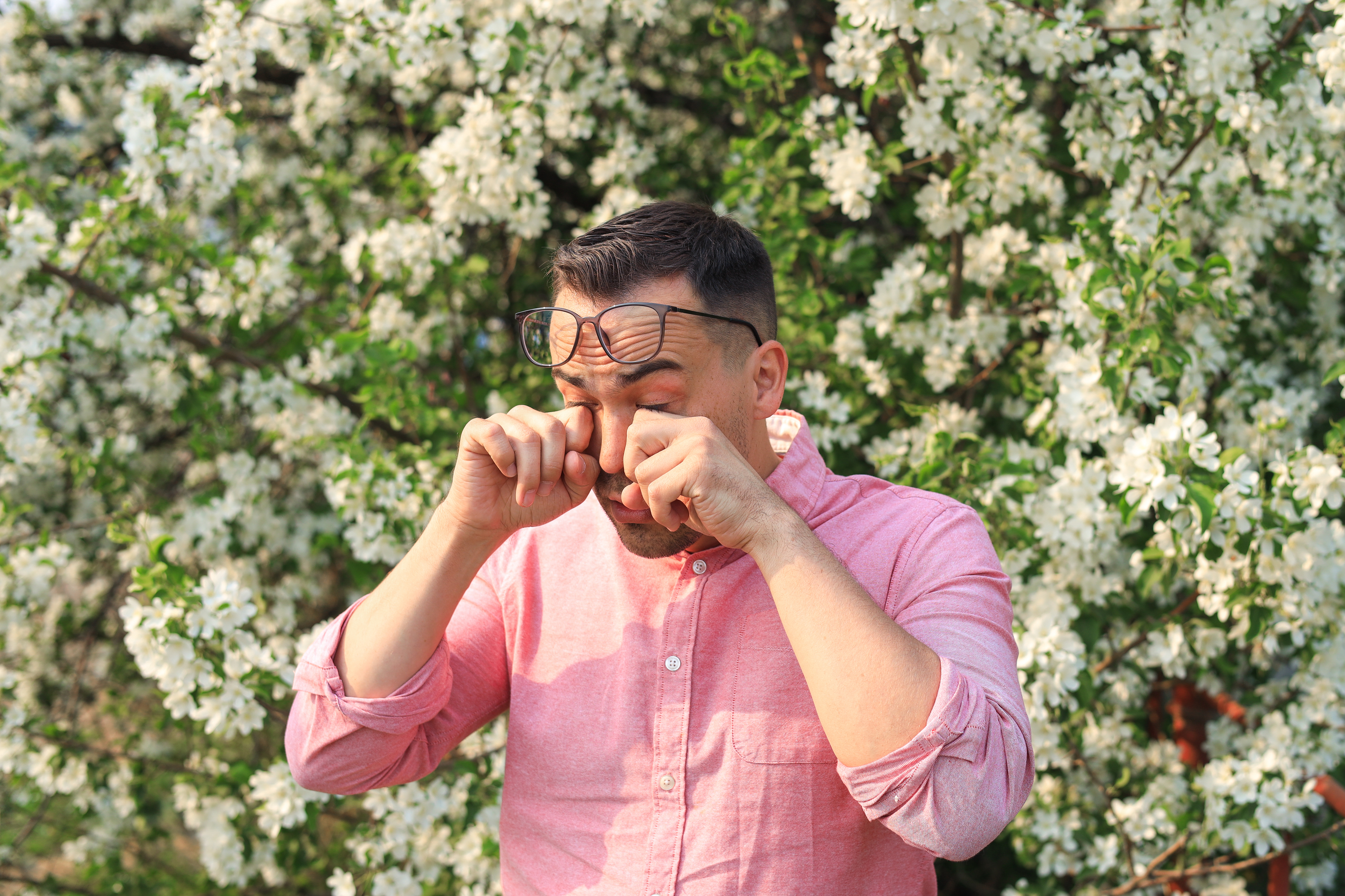 A man stands outside in front of a tree in bloom, his glasses are pushed up onto his brow while he rubs his itchy eyes.